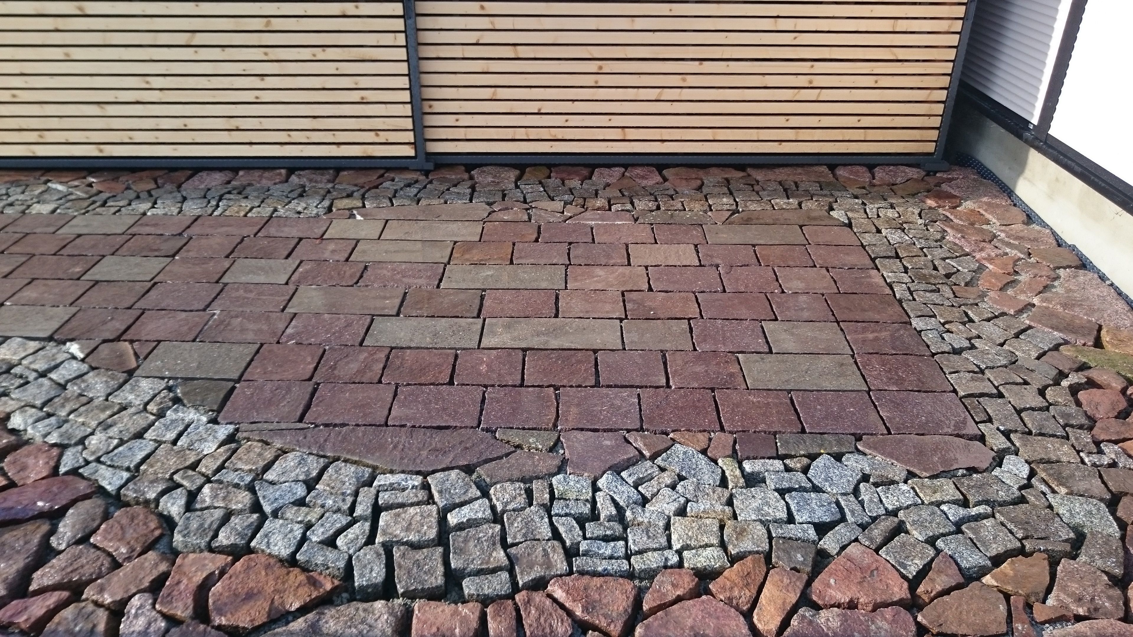 Fertige Pflasterfläche aus rotem Porphyr, eingefasst von einem Bogen aus Granit-Kleinpflaster, vor einem Sichtschutz aus Holzlamellen